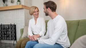 Two adults sit on a green couch in a bright living room, smiling and talking to each other.