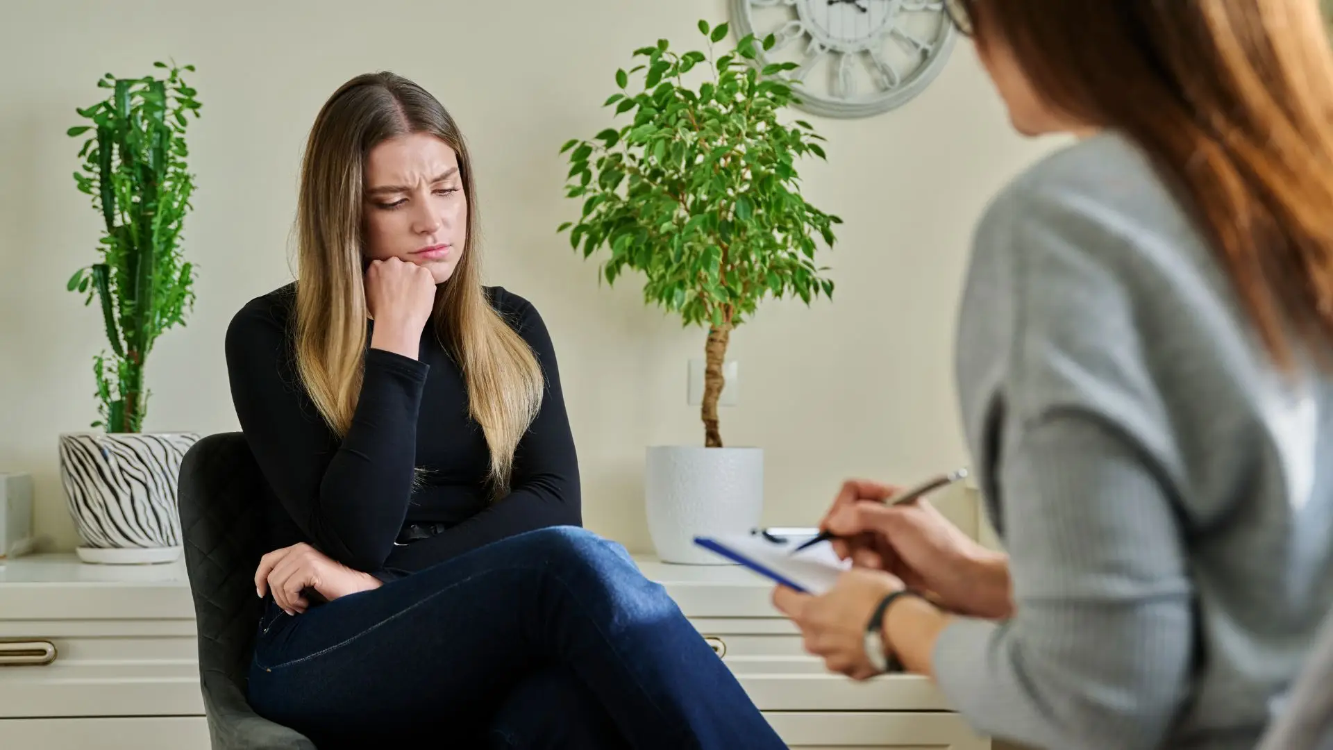 Woman in a therapy session sits pensively with a hand on her chin, while a therapist takes notes nearby in a bright room with plants in the background.